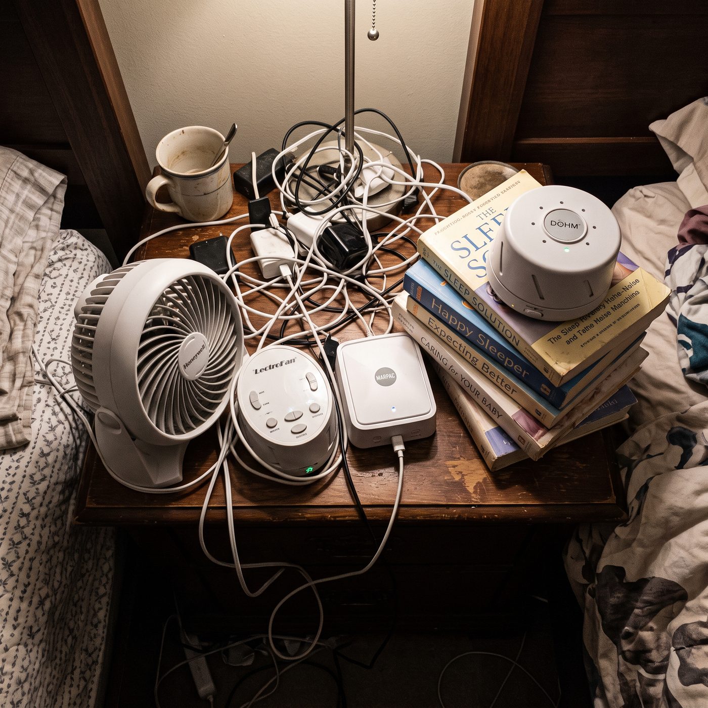 A nightstand overcrowded with multiple sound machines, a fan, and tangled cords